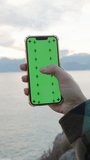 Vertical video. Against the Backdrop of Mountains and Sunset, a Woman Swipes Her Finger Across the Green Screen of a Smartphone While Standing High on a Cliff by the Sea.