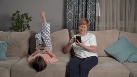 Cute girl laying on sofa in living room surfing smart phone, using app, senior woman, grandmother scrolling through social media. Resting with phone at home, smiling, laughing together. 
