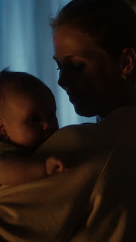 Vertical Format Portrait Video of Happy Mother Holding Adorable Newborn Baby in Cozy Dark Kids Bedroom in the Evening. Caring Mom Hugging the Neonate Toddler and They are Bonding Before Sleep.
