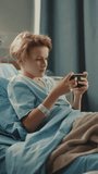 Vertical Screen. Hospital Ward: Handsome Young Boy Resting in Bed, Uses Smartphone, Plays Online Video Games on Internet with Mobile Phone. Happy Patient Recovering after Sickness