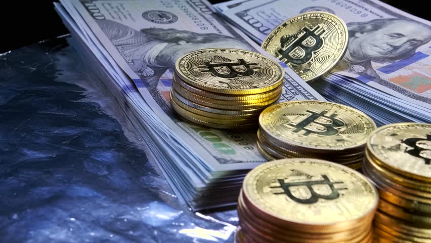 Golden physical Bitcoin coins on US dollars slow rotating money background, close up.  BTC - Bit Coin and 100 dollar bills stacks. Electronic crypto currency.