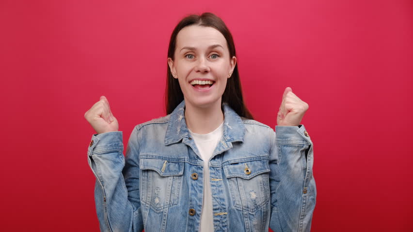 Portrait of joyful surprised young woman wearing denim jacket say wow omg what spreading hands doing winner gesture put arm on face screech, posing isolated over red color background wall in studio