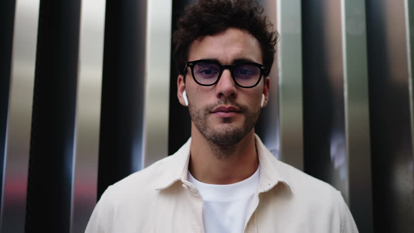 Young adult male with earbuds, possibly in his 30s stands confidently in urban setting. Caucasian handsome man in glasses dressed in casual wear look at camera. City life, modern style, individuality