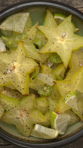 Top view of carambola or star fruit spicy salad with frozen green chili, lime pieces, lime juice, and a lot of black pepper in bowl. Table spin. Vertical video.