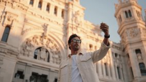 Young man travel blogger with glasses captures selfie or video in front of ornate historical building. Male tourist creating content, sharing his experience against magnificent architecture