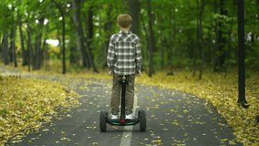 A young boy is gliding on a segway along a path in the wooded area, with trees, grass, and a natural landscape surrounding him