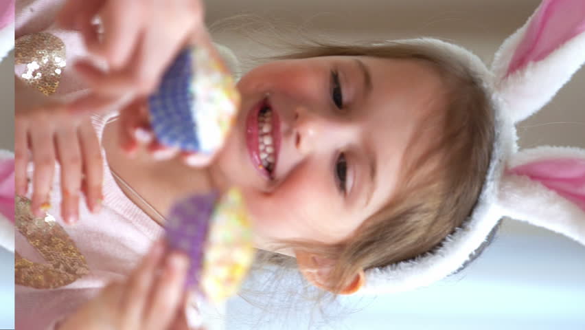 Cute little girl wearing funny rabbit ears and her young mother are cooking Easter cupcakes sitting at a festive table with basket, eggs and Bunny