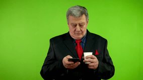 Senior businessman in suit drinking cofee and texting message on smartphone. Mature person energetically types text with fingers on phone, conducts correspondence. Green screen. Chroma key.