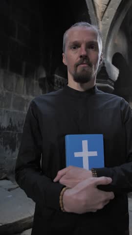 Vertical: A handsome young priest with a fashionable hairstyle blesses and forgives sins by crossing the hand of a parishioner. Caucasian stylish cleric. Religion concept. Prayer and trust in God