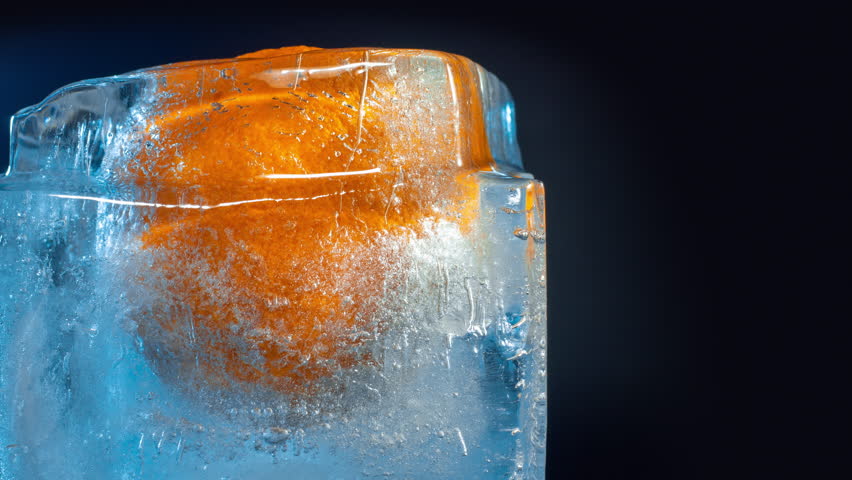Close-up view of frozen orange inside a block of ice with blue light melting against dark background. Time-lapse sequence