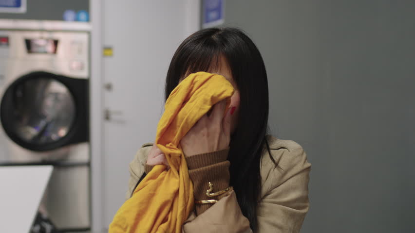 With bangs framing her face, an Asian woman takes a moment to breathe in the fresh scent of her recently laundered clothes at the laundromat, wearing a content expression. Royalty-Free Stock Footage #3444711631