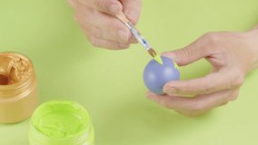 The boy's hand is holding a blue Easter egg, the other hand is holding a brush to paint cute green patterns. Scene on blue background with acrylic colored jars, video for advertising
