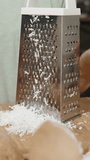 Vertical video. The Woman Grated Coconut Shavings Using a Metal Grater, and She Lifts It, Revealing the Finely Grated Coconut Flesh Falling from It.