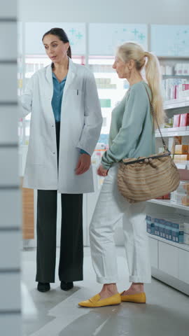 Vertical Video. Pharmacy: Professional Caucasian Female Pharmacist Helping Caucasian Senior Female Customer with Medicine Recommendation, Advice, Talking. Drugstore with Full of Health Care Products