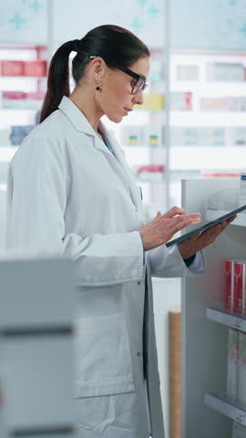 Vertical Video. Pharmacy Drugstore: Beautiful Caucasian Pharmacist Uses Digital Tablet Computer, Checks Inventory of Medicine, Drugs, Vitamins, Health Care Products on a Shelf. Professional Pharmacist