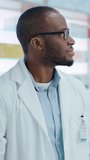 Vertical Video. Pharmacy Drugstore Checkout Counter: Portrait of Handsome Professional Black Male Pharmacist Wearing White Lab Coat, Looks at Camera, Smiles. Shelves with Medicine Packages