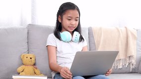Young Asian elementary girl doing homework from school at home on weekend, schoolkids student happy using technology laptop computer learning or online meeting with friends for assignment group report