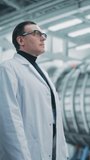 Vertical Portrait of a Professional Male Heavy Industry Engineer, Worker Wearing White Laboratory Robe and Safety Glasses. Confident Caucasian Industrial Specialist Standing in a Factory Facility