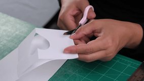 a woman cuts out a white heart shaped paper with scissors.