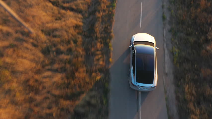 Aerial shot of electrical car driving through country road at summer evening. Modern vehicle passing rural highway. Ecology friendly car riding on electric charge along motorway. Top view.
