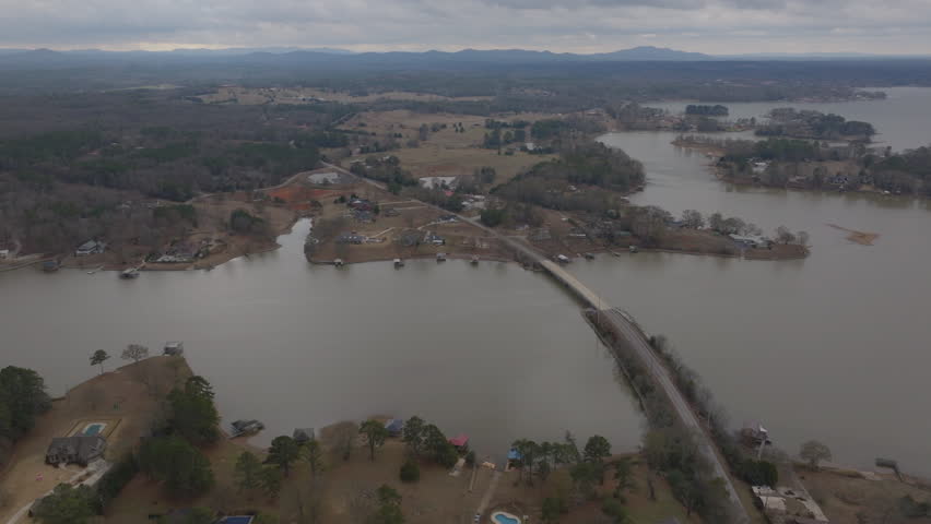 Drone footage over Lincoln, Alabama, displaying lakes, a bridge with cars, trees, and distant mountains during daytime