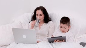 woman drinks clean water from glass cup lies sits in bed with laptop next to four-five-year-old son with tablet in hands work online training on Internet educational games spending time with son