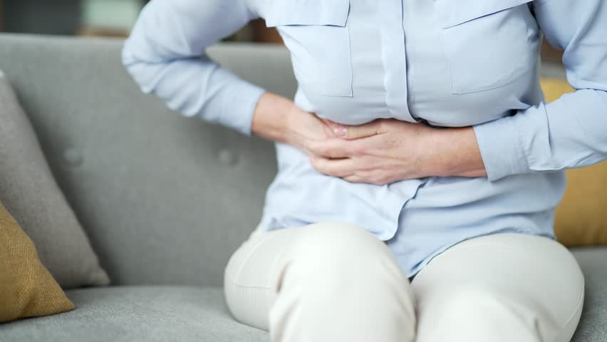 Close up. Senior female hands holding massaging right side of abdomen. Elderly woman feels sharp stomach pain sitting on sofa in living room at home. Sick adult lady suffers from liver pain or spasms