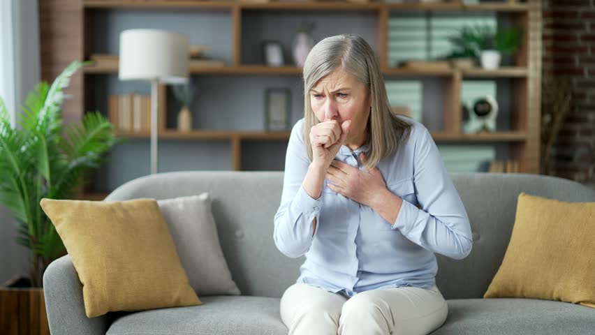 Sick senior gray haired female coughing sitting on sofa in living room at home. Sick elderly retired woman with a cold or flu feels bad, holds her hand to her chest. She has fever or bronchitis