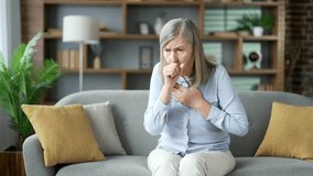 Sick senior gray haired female coughing sitting on sofa in living room at home. Sick elderly retired woman with a cold or flu feels bad, holds her hand to her chest. She has fever or bronchitis - Powered by Shutterstock - Get 15% off with code: PIKWIZARD15