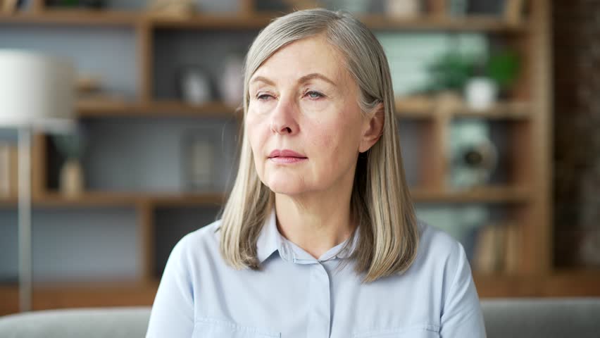 Portrait of serious senior gray haired female sitting on sofa in living room at home. Confident elderly retired woman poses looking at the camera. Headshot of a housewife in a shirt indoors. Close up Royalty-Free Stock Footage #3445090667