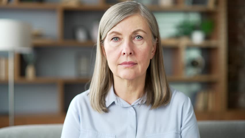 Portrait of serious senior gray haired female sitting on sofa in living room at home. Confident elderly retired woman poses looking at the camera. Headshot of a housewife in a shirt indoors. Close up