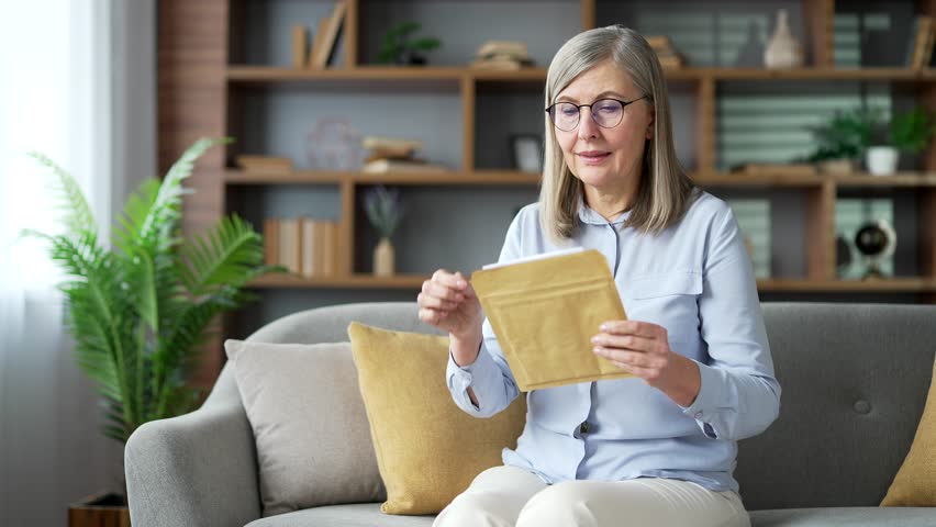 Happy senior gray haired female reading letter with great news sitting on sofa in living room at home. Joyful smiling elderly retired woman is satisfied with pleasant notification, celebrates success Royalty-Free Stock Footage #3445093293