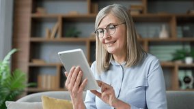 Senior woman with gray hair browsing digital tablet sitting on sofa in living room at home. Elderly female pensioner reads, writes messages, chats, does online shopping, uses an application. Close up - Powered by Shutterstock - Get 15% off with code: PIKWIZARD15