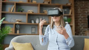 Senior gray haired female using VR glasses for video call and chatting in virtual reality simulator sitting on sofa in living room at home. Elderly woman communicates remotely, has an online meeting