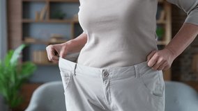 Close up of mature woman's hands trying on her big pants after losing weight. A middle aged female is happy with the success of a good figure waist after a diet while standing in living room at home - Powered by Shutterstock - Get 15% off with code: PIKWIZARD15