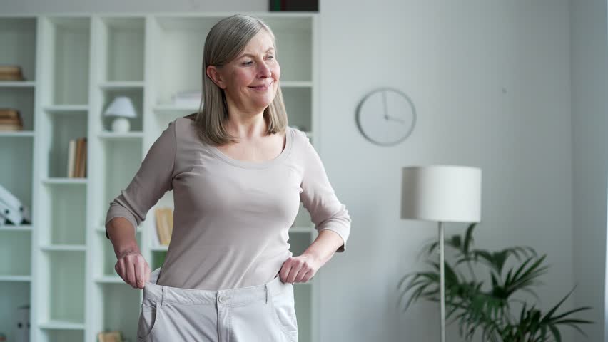 Happy senior gray haired female feels joy after losing weight. A smiling, satisfied middle aged woman tries on her big pants, which she wore before the diet while standing in the living room at home