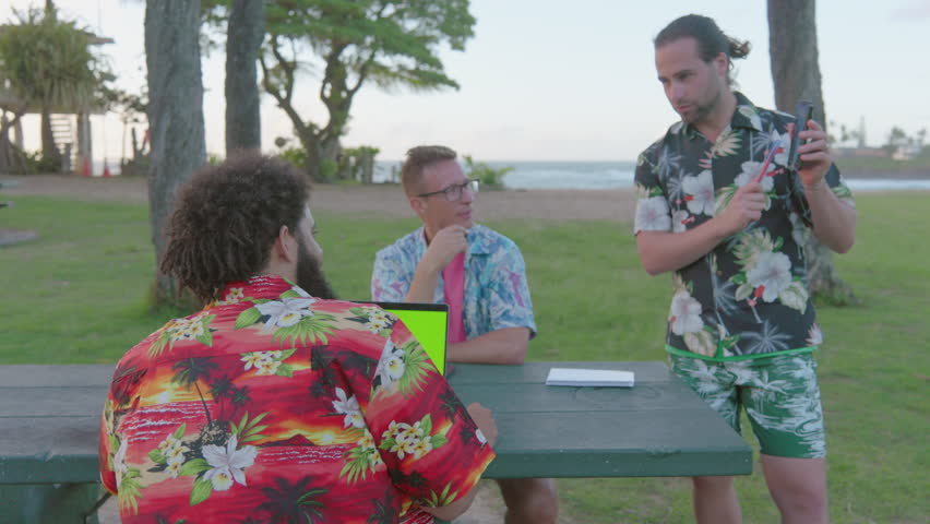 3 young men wearing Hawaiian shirts enjoy a picnic and working remotely, surrounded by trees, grass, vibrant plant life, while donning hats and engaging in recreational activities. Working remotely  Royalty-Free Stock Footage #3445407425