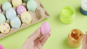 Hand model is holding an egg in pink color and using a paintbrush to paint it golden. Some paint jars decorated with an egg carton of colorful Easter eggs. Easter Day concept