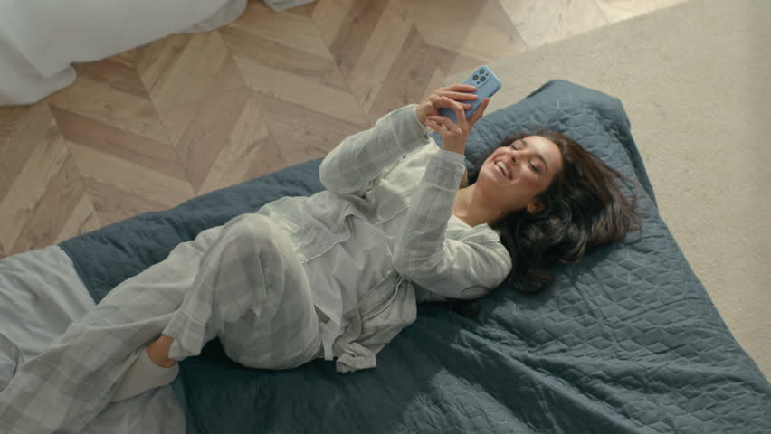 Top view young cheerful brunette woman in pajamas lying on bed cozy comfortable home inside bedroom female roll over chatting texting smartphone mobile phone telephone communicate smiling weekend joy