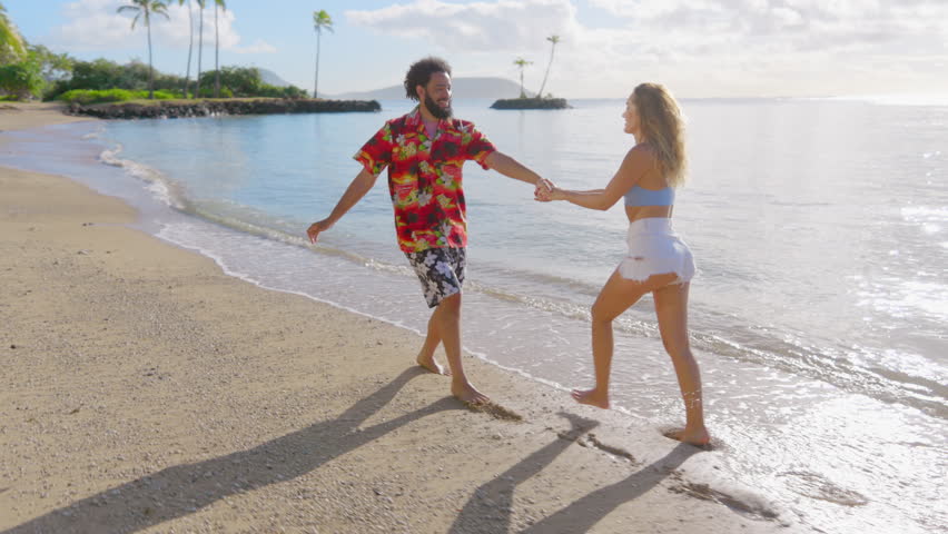 A happy young couple holding hands on the beach, running and enjoying the water, sky, and horizon while surrounded by palm trees, creating a gesture of love and leisure. Royalty-Free Stock Footage #3445590821