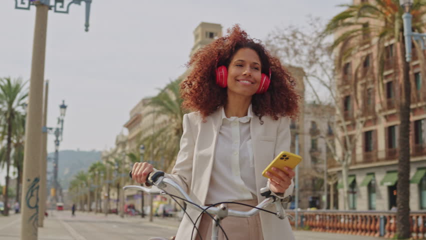 latin woman wallking with a bike, headphones and smartphone, in barcelona tourist area