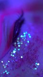 Extreme closeup view of woman blinks, tear flows from eye, female cries. Eye with makeup with sequins, arrows. Cinematic shot illuminated neon lighting. Selective focus, handheld. Vertical video shot