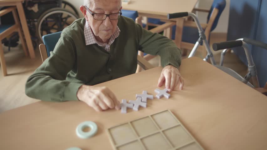 an elderly man carefully places a board game tile in a retirement home, engaging in leisure activity that promotes mental stimulation, social interaction, and a sense of community among residents Royalty-Free Stock Footage #3445704285