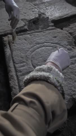 Vertical: Hands in gloves, two archaeologists using brushes sweep away dust from the diaracal sign of Libra carved on stone in an ancient temple. Astrological ancient symbols, alchemy designations