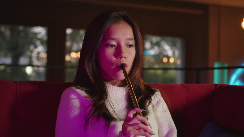 Asian young woman takes of mouthpiece to her lips, inhales and blows out puffs of white smoke at shisha cafe. Pretty adult girl smoking hookah sitting at couch at bar. Female smoker. Front view