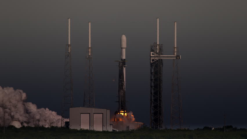 A rocket lifts off at dusk in slow-motion from launch pad in Florida as it flies into space with a satellite as cargo. Excellent view of smoke and flames at launch pad.