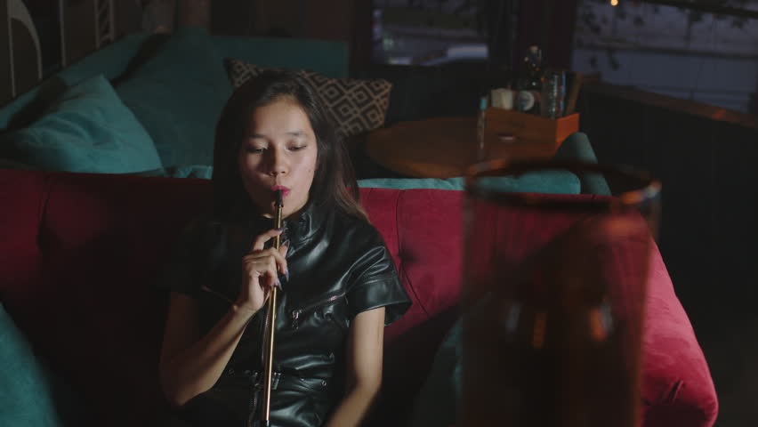 Asian young woman takes of mouthpiece to her lips, inhales and blows out puffs of white smoke at shisha cafe. Pretty adult girl smoking hookah sitting at couch at bar. Female smoker, bad habit