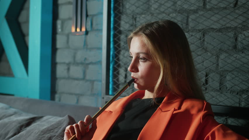 Blonde young woman takes of mouthpiece to her lips, inhales and blows out puffs of white smoke at shisha cafe. Pretty adult girl smoking hookah sitting at couch at bar. Female smoker, bad habit