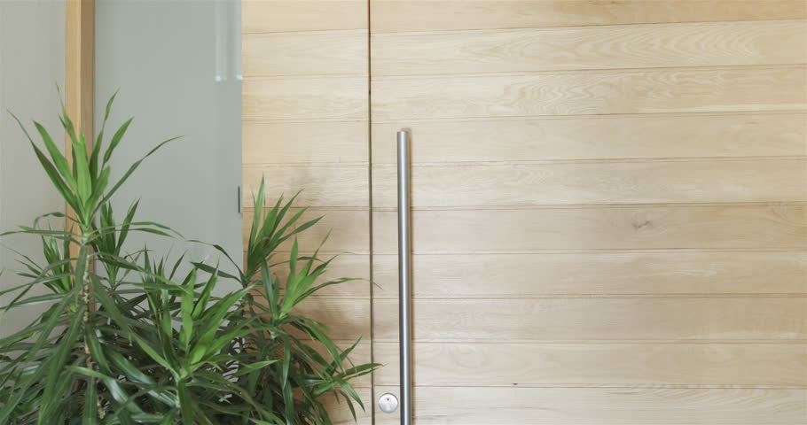 Young Asian man stands smiling at the entrance of a modern home. Welcoming atmosphere is evident as he opens the door to guests or family, slow motion.