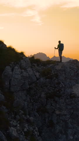 AERIAL, SILHOUETTE, LENS FLARE: Stunning sunset illuminates the Alps and hiker standing on edge of a cliff and observing the picturesque mountain landscape. Man watching the sunrise in the mountains.